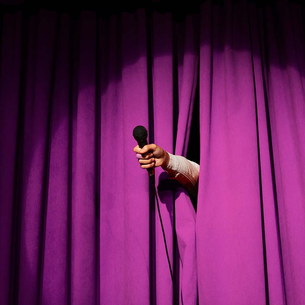 A hand holding a microphone reaches out behind closed purple theater curtains